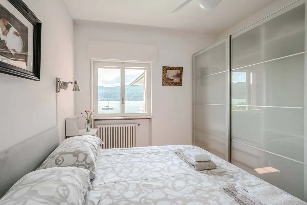 Un dormitorio con cama, ventana, radiador, mesita de noche con lámpara y un cuadro enmarcado en la pared.