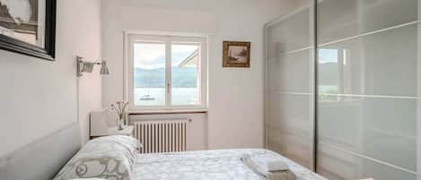 Une chambre à coucher avec un lit, une fenêtre, un radiateur, une table de chevet avec une lampe et un tableau encadré au mur.