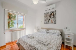 A bedroom with a bed, a nightstand, a window with a view, and an air conditioning unit. - Italian Lakes Lakeside Villa With Direct Lake Access (Ranco)