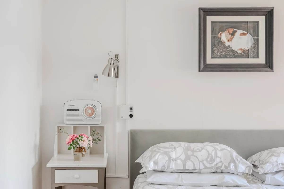 A bedroom with a bed, a bedside table with a radio, a framed picture on the wall, and a small vase with flowers.
