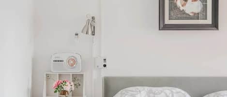 A bedroom with a bed, a bedside table with a radio, a framed picture on the wall, and a small vase with flowers.