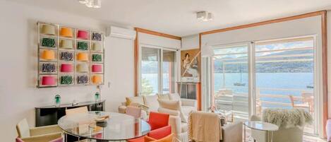 A modern living room with a glass table, colorful chairs, a sofa, and a view of the sea.