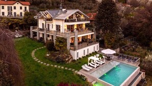 Une grande maison à plusieurs étages, dotée d’une piscine, entourée d’une végétation luxuriante et de collines.