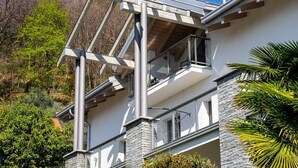 Une maison moderne à deux étages, dotée d’un balcon, d’une façade en pierre et offrant une vue dégagée sur les arbres environnants.