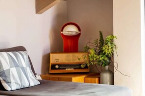 Ein Bett mit Kissen, ein Nachttisch aus Holz mit einem Radio, eine rote Lampe und eine Vase mit Grünpflanzen.