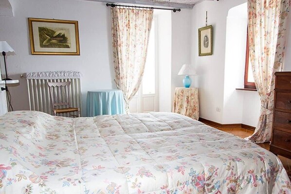 A bedroom with a floral bedspread, a wooden dresser, a chair, and a table with a lamp. - Stylish Lake Maggiore Apartment With Lake Views (Maccagno con Pino e Veddasca)