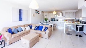 A modern living room with a beige sectional sofa, a coffee table, and a dining area with a white table and bar stools. The kitchen in the background has white cabinets and a central island. - Dumenza Villas (Dumenza)