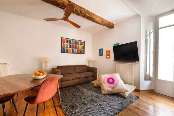 A living room with a wooden ceiling beam, a brown sofa, a round dining table with chairs, a fruit bowl, a television, and a patterned rug. - Lesa Apartments (Lesa)