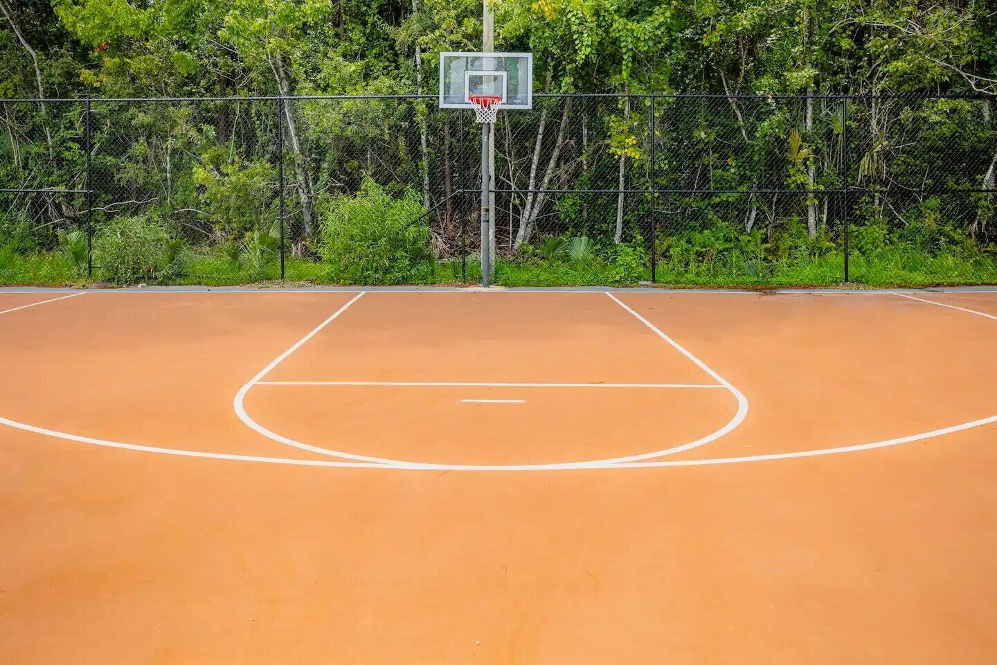 En udendørs basketballbane med hegn og træer i baggrunden.