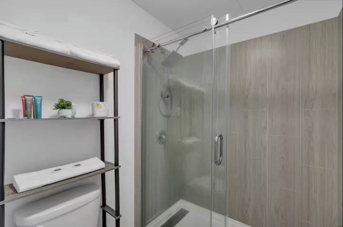 A modern bathroom with a glass shower enclosure, a white shelf with toiletries, and a wooden vanity.