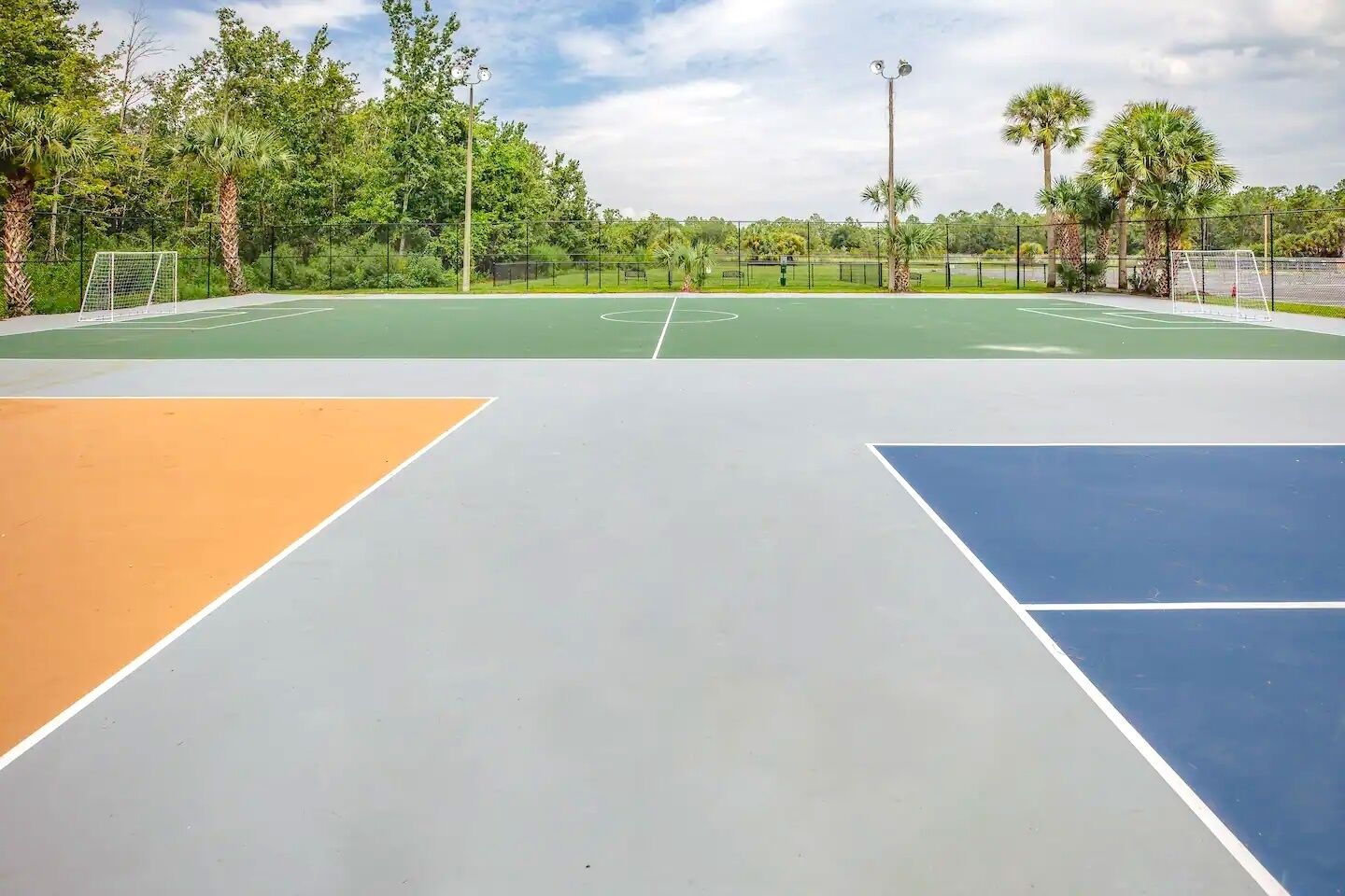 Una cancha de baloncesto al aire libre con superficie de juego verde, naranja y azul, rodeada de árboles y una valla.