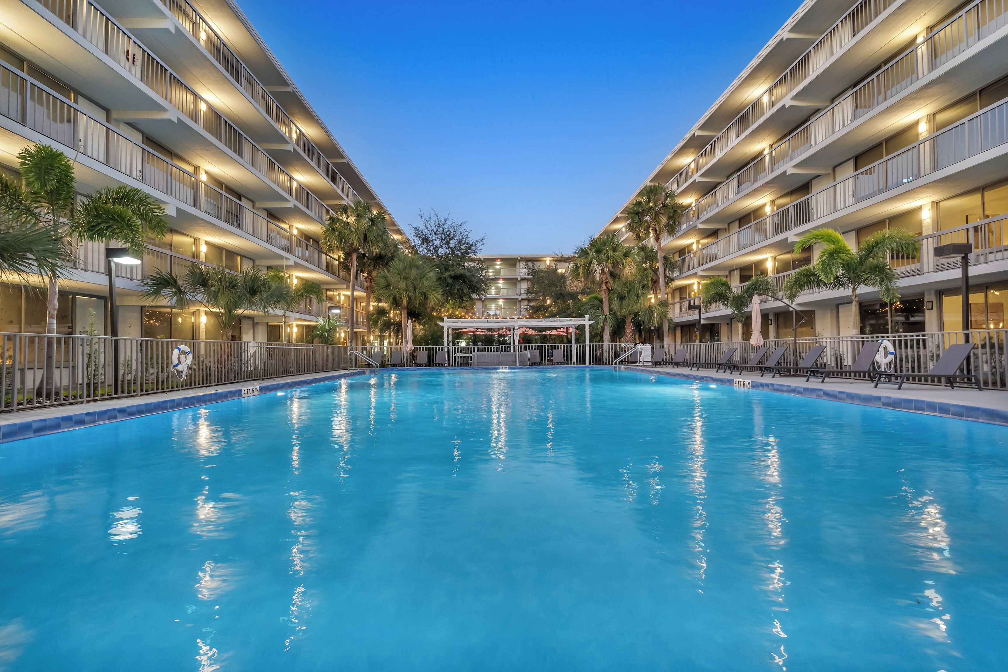A large indoor swimming pool with multiple balconies, palm trees, and a covered area in the background.