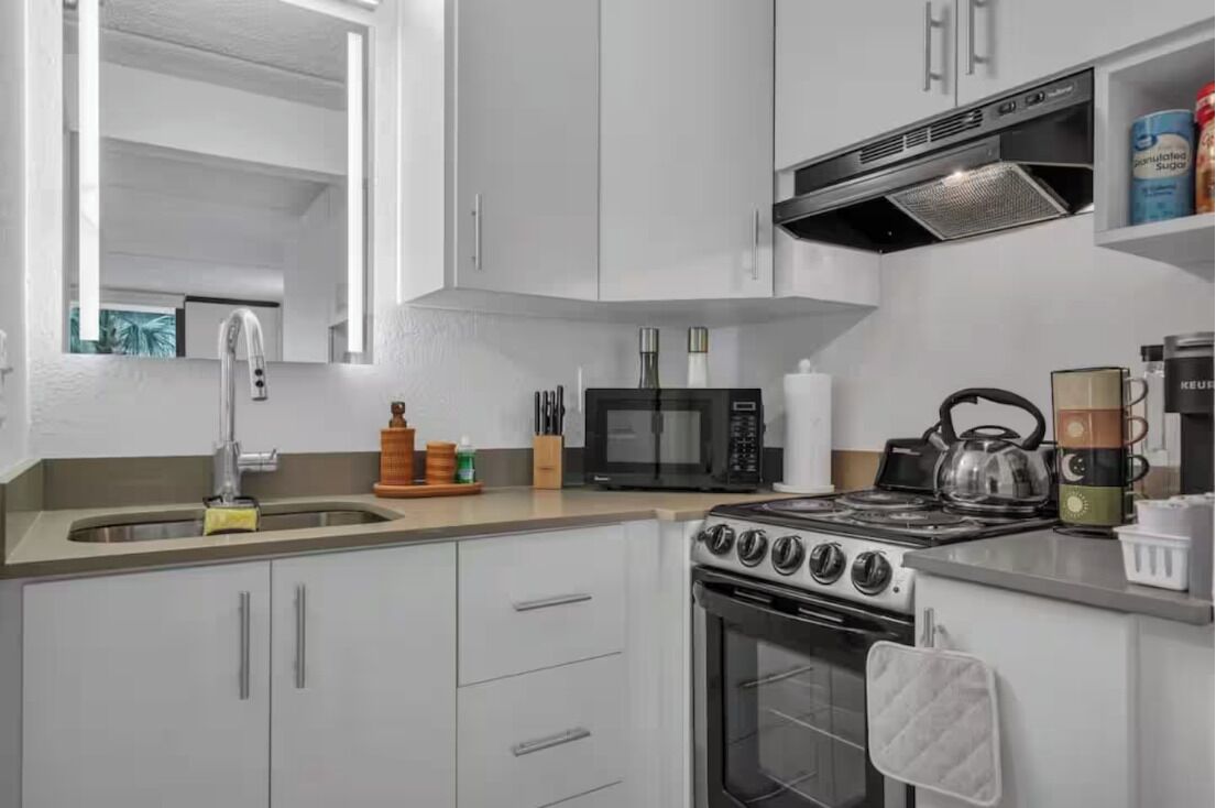 A modern kitchen with white cabinets, a stainless steel sink, a microwave, a stove, and a kettle.