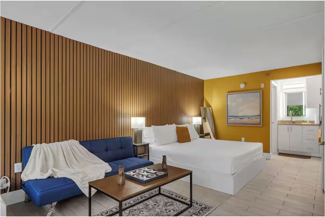 Une chambre d’hôtel moderne avec un mur en lambris de bois, un canapé bleu, un lit blanc et une petite kitchenette.