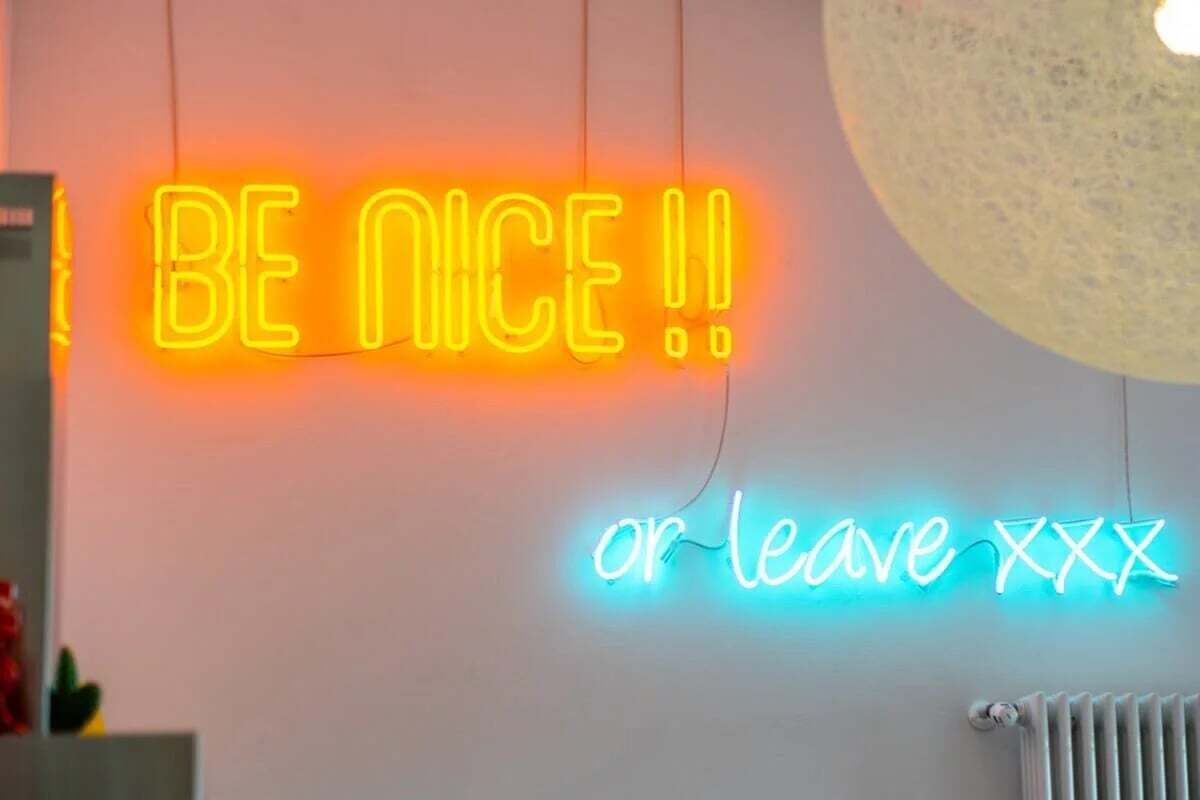 Un cartello luminoso al neon con il testo 'BE NICE!!' e 'or leave XXX' su un muro.