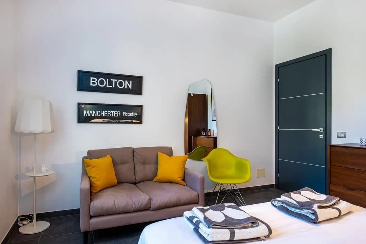 A modern hotel room with a sofa, two chairs, a bed, a mirror, and two framed signs on the wall.