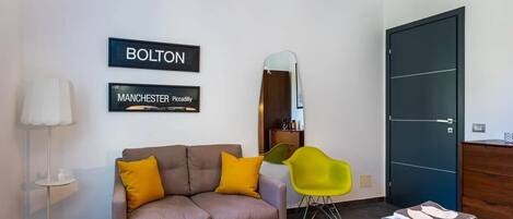 A modern hotel room with a sofa, two chairs, a bed, a mirror, and two framed signs on the wall.