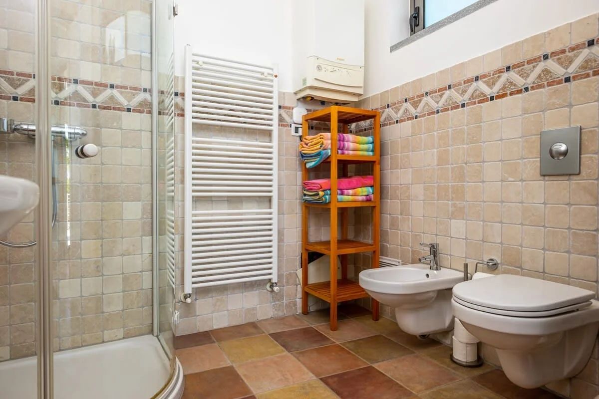 A bathroom with a glass shower enclosure, a white radiator, a wooden shelf with towels, two bidets, and a toilet.