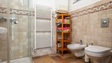 A bathroom with a glass shower enclosure, a white radiator, a wooden shelf with towels, two bidets, and a toilet.
