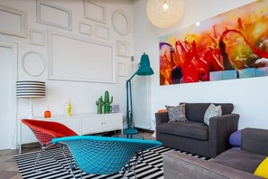 Ein modernes Wohnzimmer mit geometrischer Wandgestaltung, einem farbenfrohen abstrakten Gemälde, einem grauen Sofa mit gemusterten Kissen, einem roten Sessel und einem blauen Stuhl mit Metallgestell.