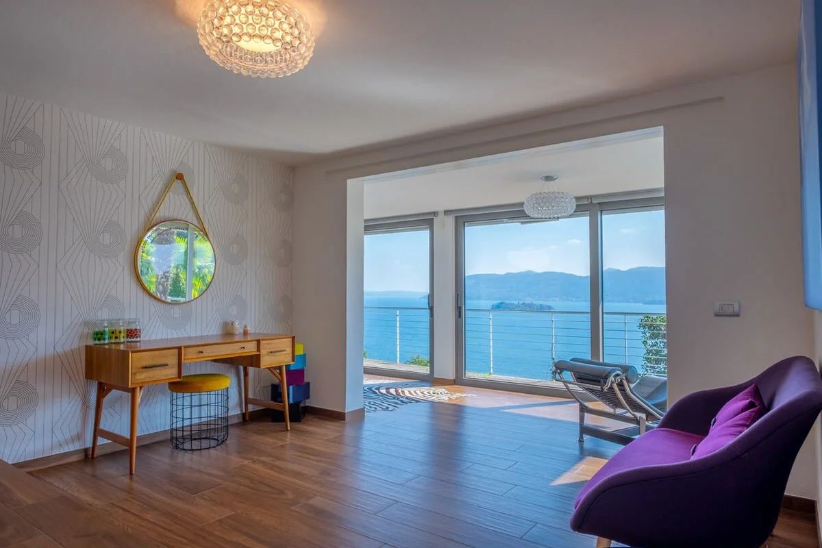 A room with a wooden desk, a purple chair, a round mirror, and a view of the sea through large windows.