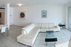 Ein modernes Wohnzimmer mit einem weißen Ecksofa, einem Glastisch und einem weißen Esstisch mit Stühlen.