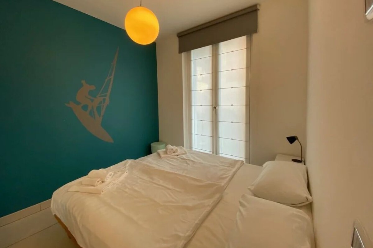 Une chambre à coucher avec un lit, une fenêtre avec des stores, une lampe fixée au mur et un élément décoratif mural.