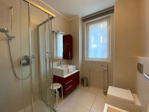 A modern bathroom with a glass shower enclosure, a red vanity, a white sink, and a large window. - Maccagno Apartments (Maccagno con Pino e Veddasca)