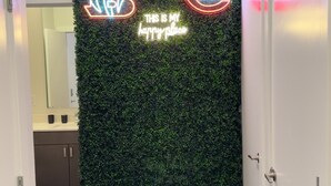 A hallway with a green artificial hedge, neon signs for 'B' and 'Reds', and a 'This is my happy place' sign. - The 513 Flat! Pool & River View! Free Gated Parking! Walk to Banks & Stadiums! (Cincinnati)
