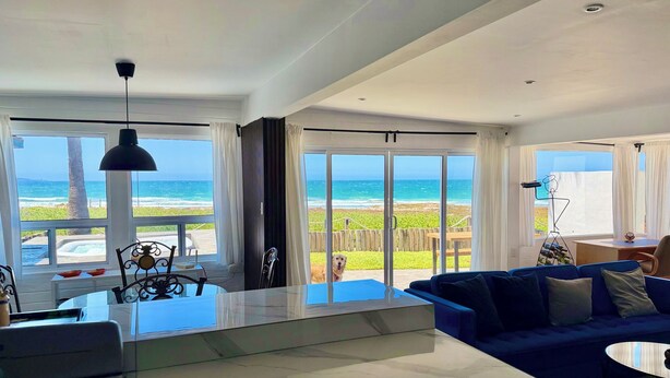 A modern living room with a large window overlooking the ocean, a blue sofa, and a kitchen area with a marble countertop. - Casa Azul - beachfront jacuzzi & sunsets (Ensenada)