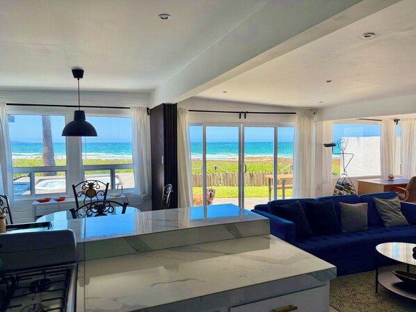 A modern living room with a large window overlooking the ocean, a blue sofa, and a kitchen area with a marble countertop.