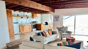Een ruime woonkamer met een bank en fauteuils, een keuken op de achtergrond, en een vloer van stenen tegels.