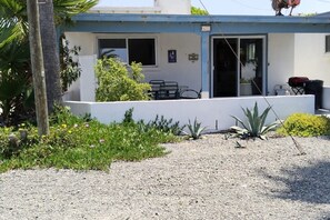Un petit bâtiment à l’extérieur bleu, avec une porte et une fenêtre. Un petit jardin avec différentes plantes et une zone recouverte de gravier se trouvent devant.