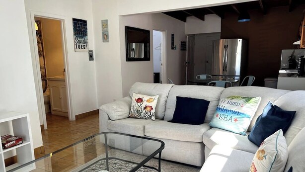 Una sala de estar con un sofá, una mesa de centro de vidrio y una cocina al fondo.
