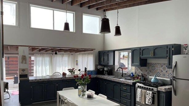 Une cuisine moderne avec des meubles de couleur sombre, une table à manger blanche et des luminaires suspendus.