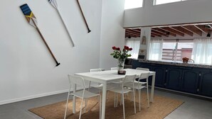 Un coin repas avec une table et des chaises blanches, un tapis tissé et une cuisine en arrière-plan.