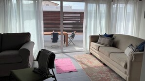Un salon comprenant un canapé, un fauteuil, une petite table et une vue, par une porte vitrée, sur un espace extérieur aménagé avec une table et des chaises.