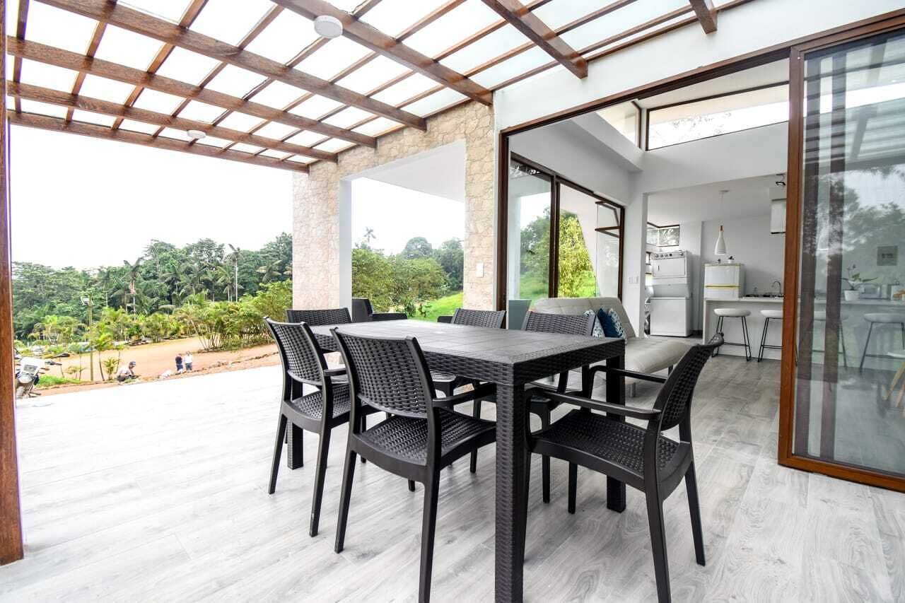 A covered patio with a dining table and chairs, a kitchen area, and a view of the surrounding greenery.