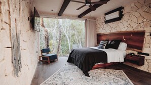 Ein Schlafzimmer mit einem großen Bett, einem Holz-Kopfschild, einem Nachttisch, einem Sessel und Blick auf Bäume durch ein Fenster.