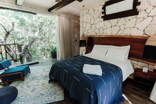 Un dormitorio con pared de piedra, una cama grande con ropa blanca, un tapete con estampado azul y blanco, y vistas a árboles a través de un ventanal.