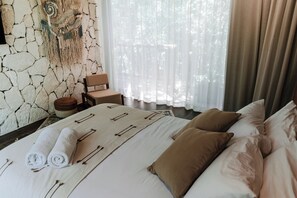 A hotel room with a stone wall, a bed with white linens and brown pillows, a small wooden chair, and a large window with sheer curtains. - Tulum Jungle Villa Aire (Tulum)