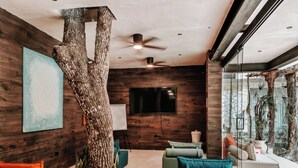A modern living room with a large tree trunk centerpiece, wooden walls, a ceiling fan, and a large window with a view of greenery. - Tulum Jungle Villa Aire (Tulum)