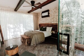 A bedroom with a bed, wooden headboard, and a view of a tropical garden through a glass door.