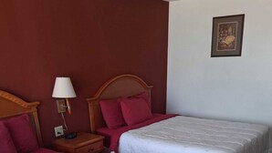 Habitación de hotel con dos camas, cada una con ropa de cama blanca y almohadas rojas. Cuenta con un cabecero de madera, una mesita de noche con una lámpara y un cuadro en la pared.