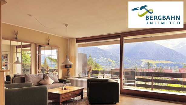 A modern living room with a large window offering a mountain view, a sofa, a coffee table, and a lamp. - Alpenjuwel Oberstdorf – Mountain Views, Balcony & Sauna, Lift Pass, Dog Friendly (Oberstdorf)