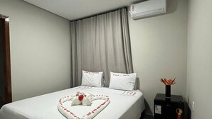Un lit bien fait, avec du linge de lit blanc et une disposition de serviettes en forme de cœur, un radiateur noir posé sur le sol et un climatiseur fixé au mur.