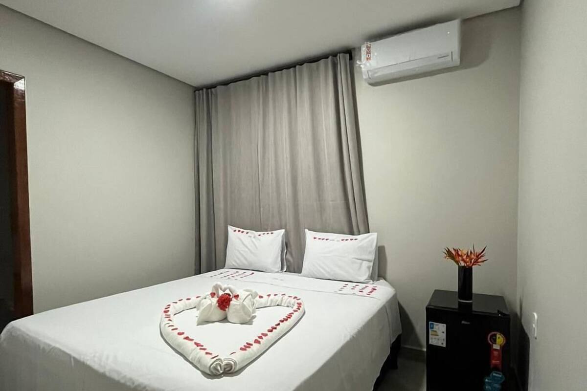 A neatly made bed with white linens and a heart-shaped towel arrangement, a black heater on the floor, and an air conditioning unit on the wall.