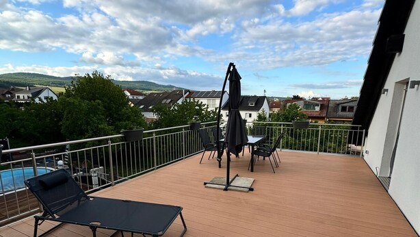 A wooden deck with a lounge chair and a table, overlooking a residential area with houses and a hill in the distance. - Cozy vacation apartment in Winkel in the heart of the Rheingau. (Oestrich-Winkel)