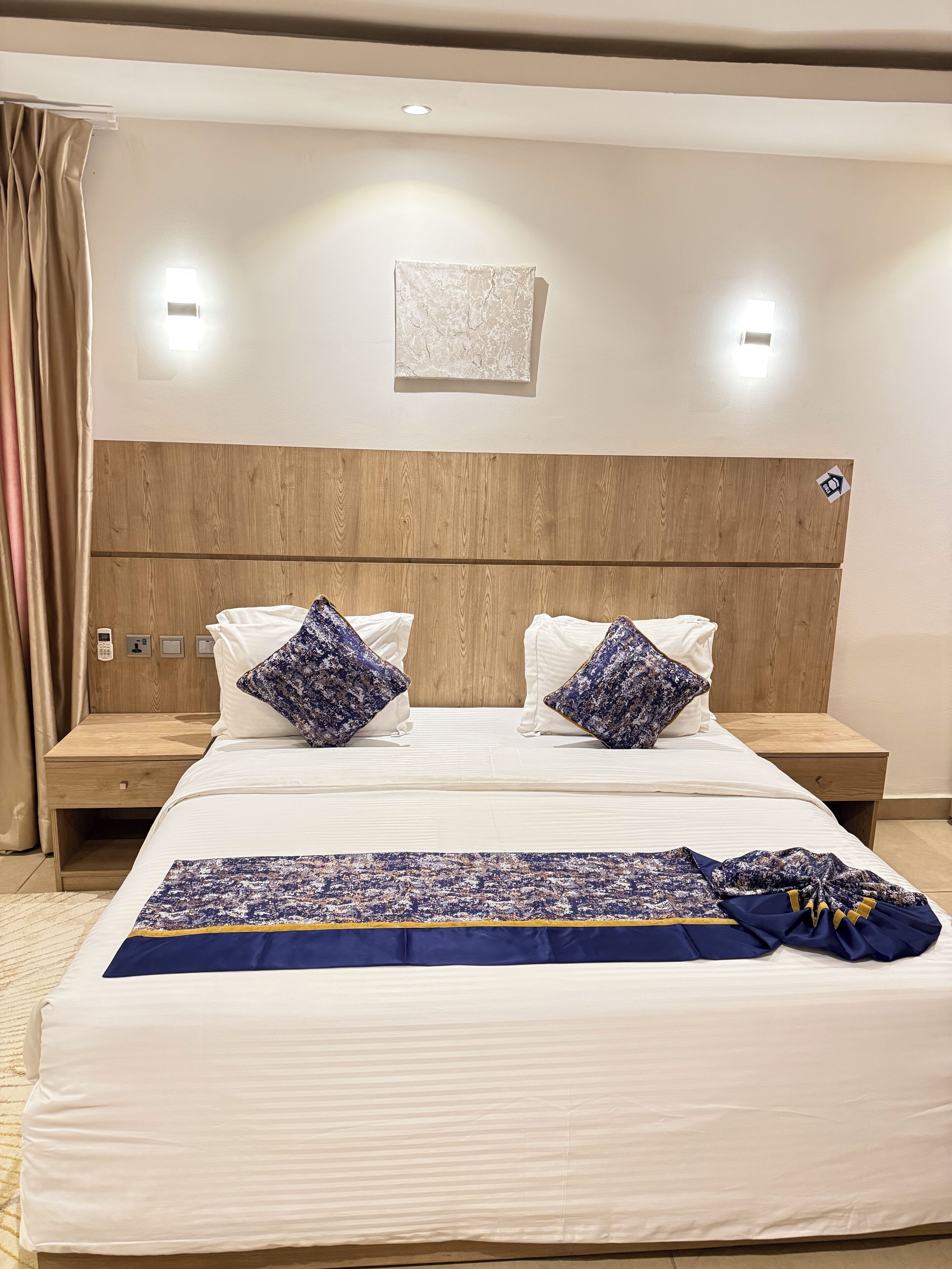 Une chambre d’hôtel avec une tête de lit en bois, deux tables de chevet, un lit avec une literie blanche et des oreillers à motifs bleus, ainsi qu’une couverture bleue et jaune.