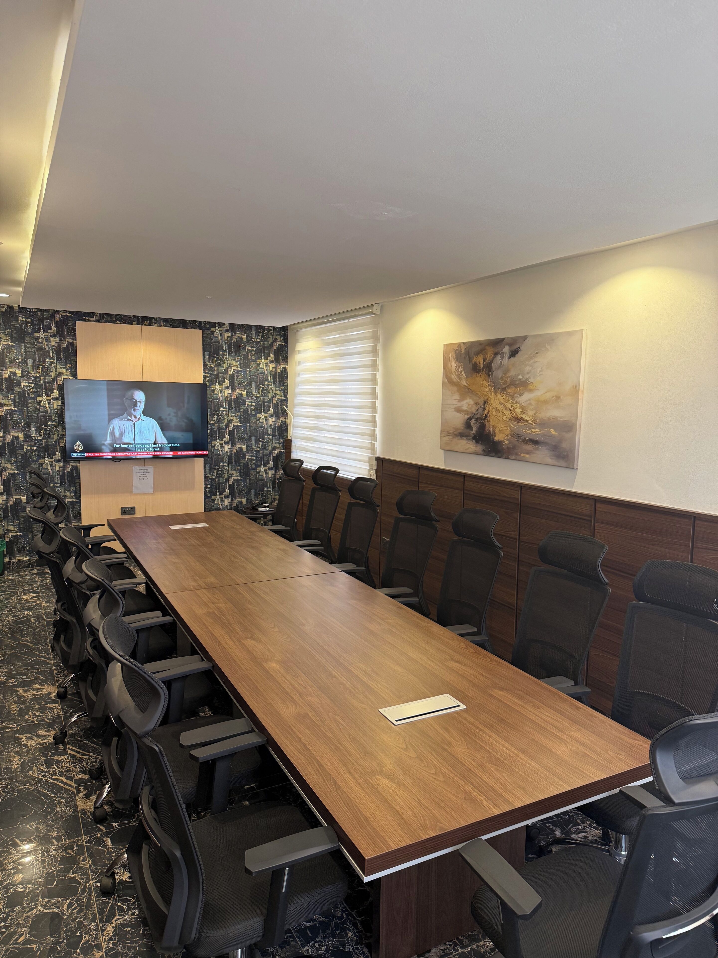 A conference room with a long wooden table, black chairs, a television on the wall, and a cityscape wallpaper.